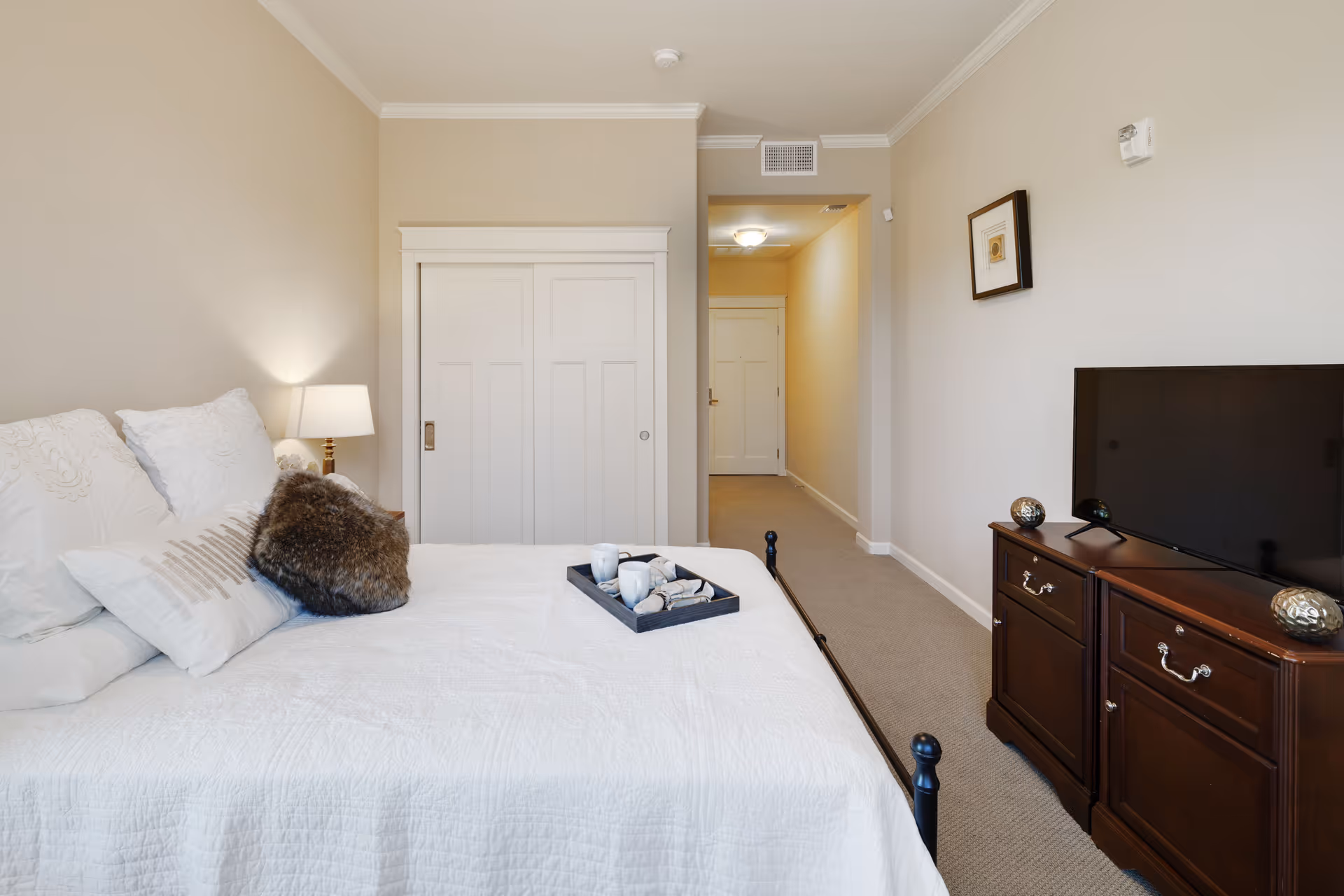 A neatly arranged bedroom with a white bedspread, decorative pillows including a furry one, and a tray with cups on the bed. There is a bedside lamp on a small table next to the bed. Across from the bed is a dark wooden dresser with a flat-screen TV on top. The walls are beige, and there is a framed picture hanging above the dresser. A hallway with a closed door at the end is visible beyond a sliding closet door.
