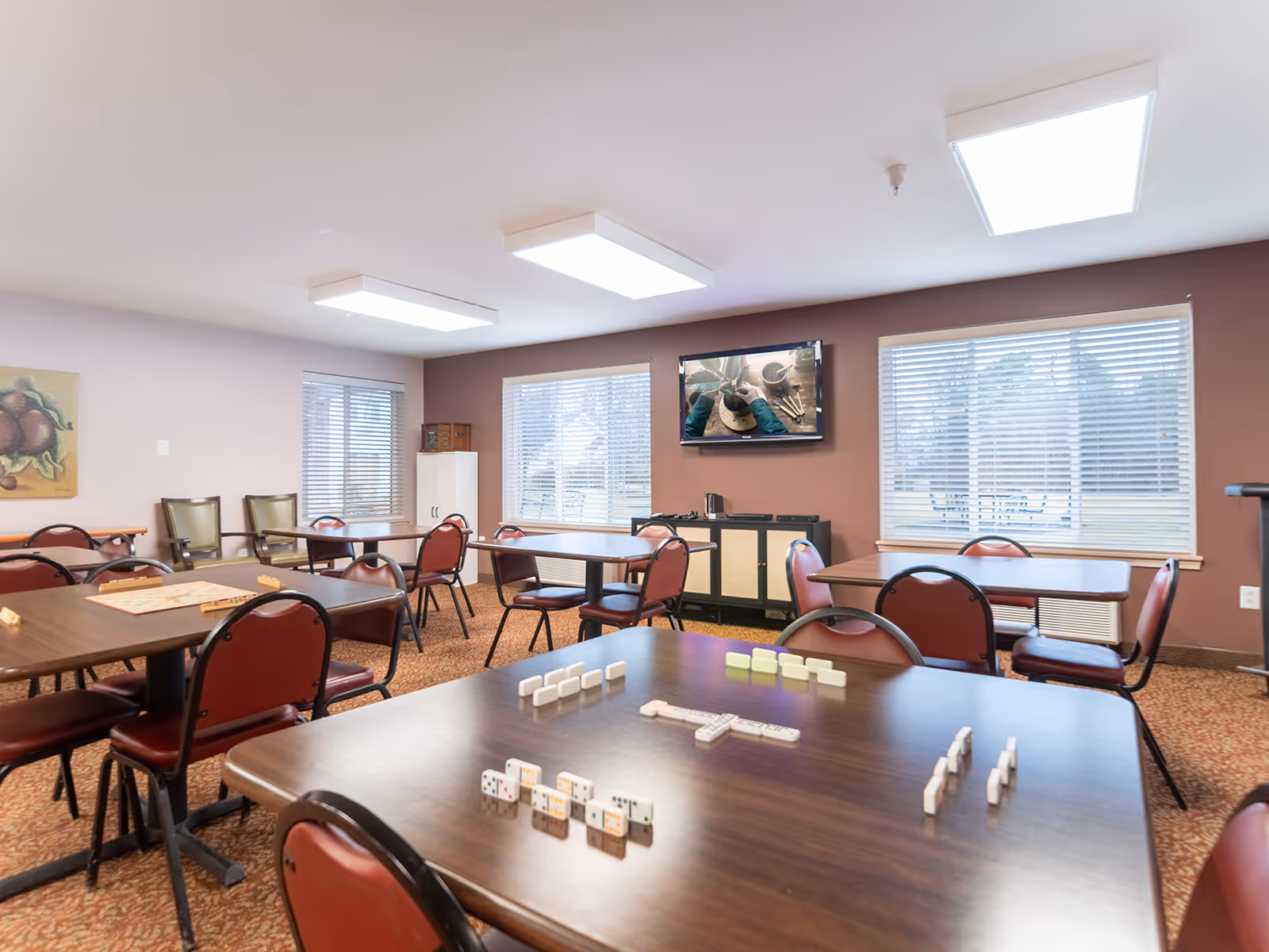 A well-lit activity room with several tables and red cushioned chairs arranged around them. Dominoes and a Scrabble board are set up on the tables, suggesting games in progress. The room has large windows with blinds, a wall-mounted TV displaying a close-up of hands working on a craft, and a cabinet against a brown accent wall. The carpet is patterned, and the ceiling has rectangular fluorescent lights.