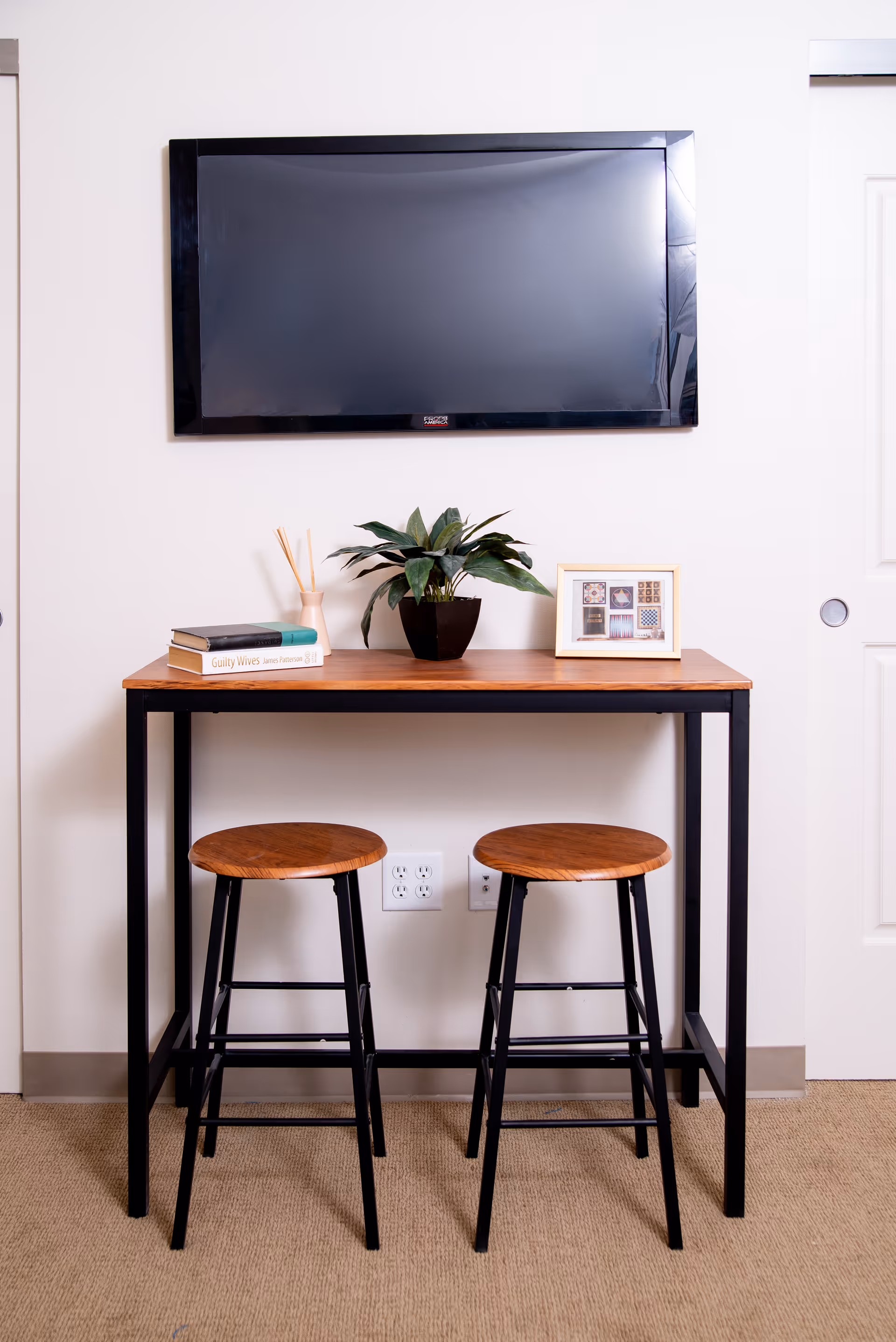 A small wooden table with black metal legs against a white wall, with two wooden stools in front. On the table are two books, a small potted plant, a reed diffuser, and a framed picture. Above the table is a mounted flat-screen TV. There is a white door to the right of the table.