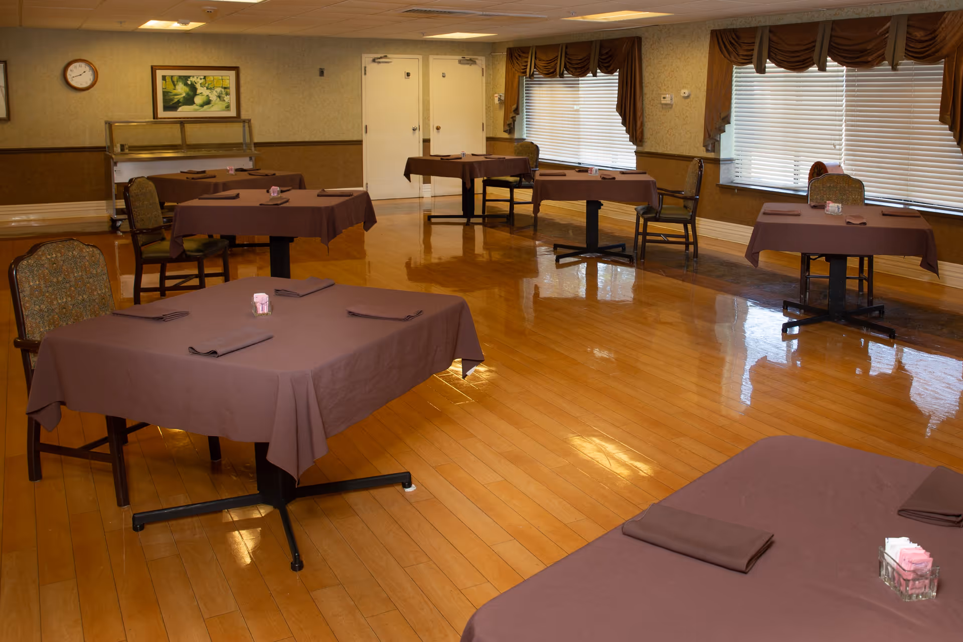 A dining room with several tables covered in brown tablecloths, each set with folded napkins and small condiment containers. The room has wooden flooring, chairs with patterned upholstery, large windows with blinds and brown valances, and a clock and framed artwork on the wall.