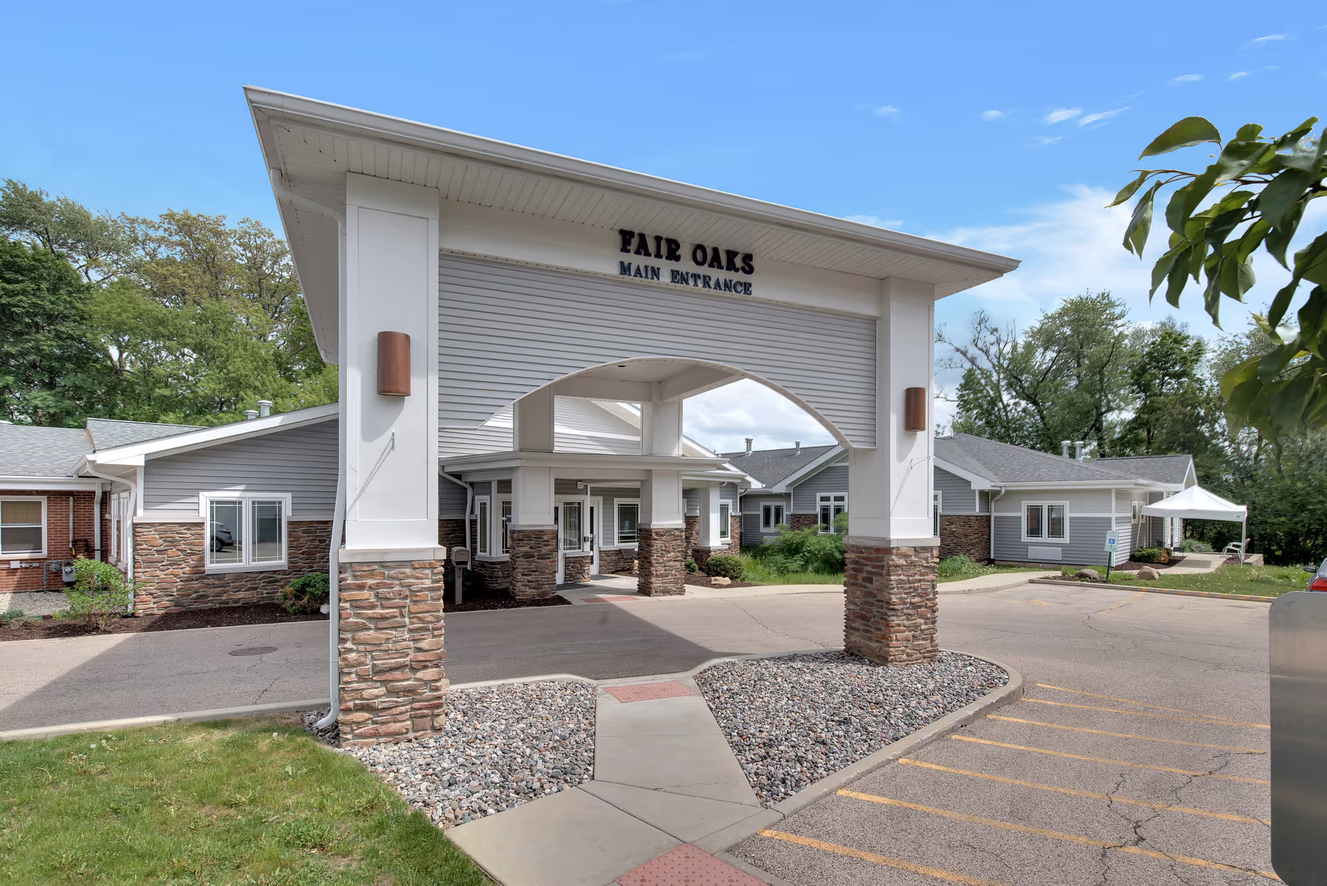 Main entrance of Fair Oaks Health Care Center featuring a covered drop-off area with stone pillars and a paved driveway surrounded by greenery and trees under a clear blue sky.