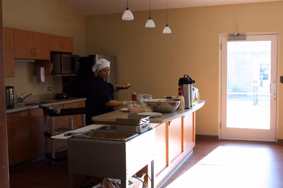 A staff member wearing a chef's hat serves food behind a kitchen island in a bright facility kitchen.