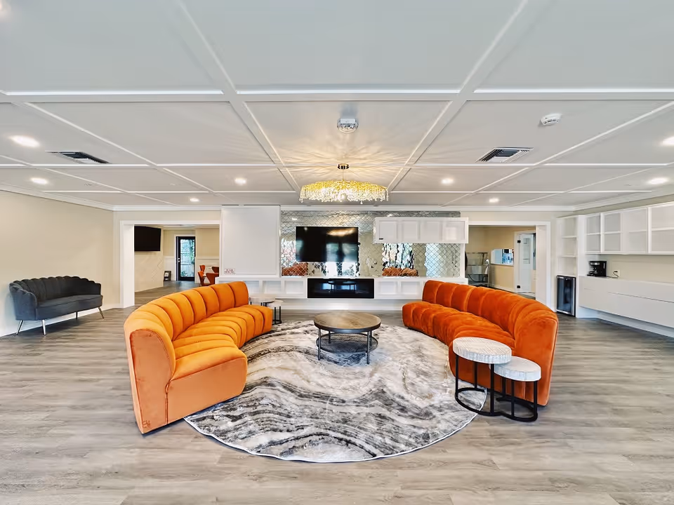 Spacious modern communal lounge with two curved orange sofas around a round rug and central coffee table facing a wall-mounted TV.