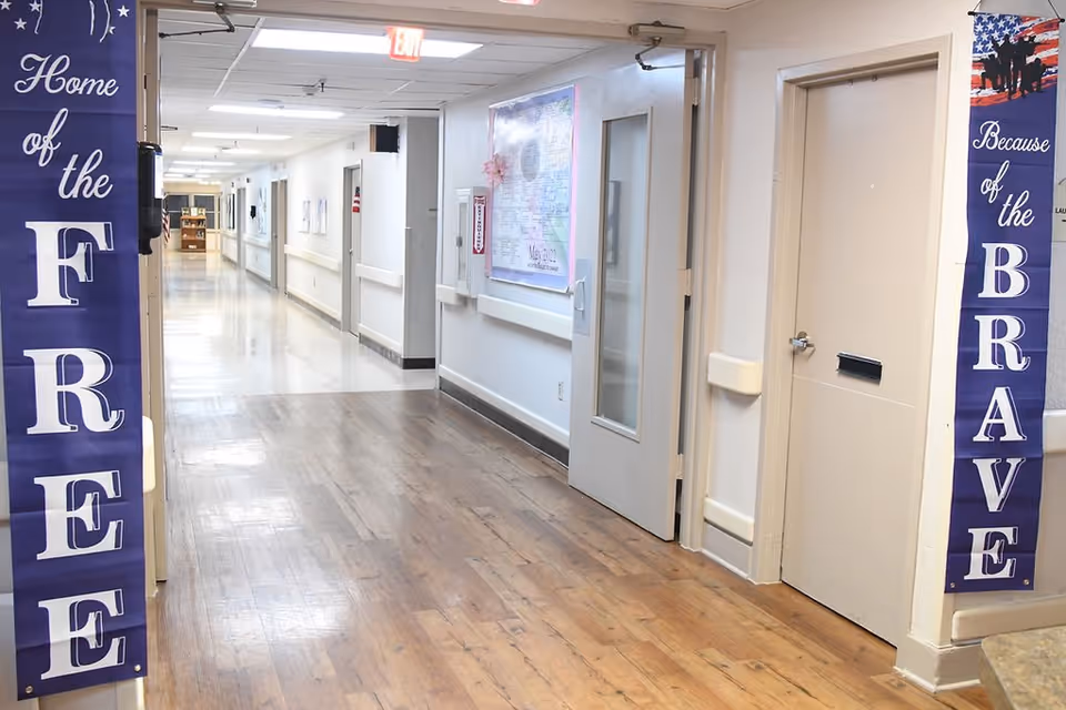 A clean, well-lit hallway in a nursing and rehabilitation facility with wood-patterned flooring and white walls. On the left side, there is a purple banner that reads 'Home of the FREE' and on the right side, another purple banner reads 'Because of the BRAVE'. Doors and handrails line the hallway, and an exit sign is visible at the far end.