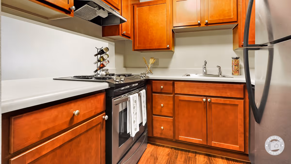 A compact kitchen with wooden cabinets, a black stove with two white towels hanging on the handle, a stainless steel refrigerator, a sink with a faucet, and a wine bottle holder on the countertop. The floor is wooden and the countertops are white.