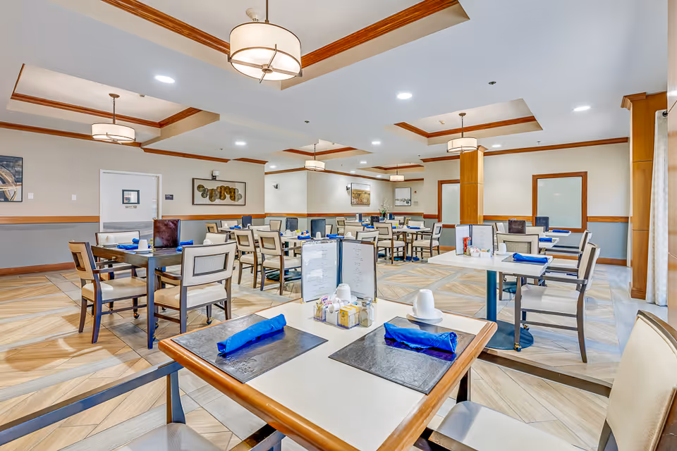 A bright and spacious dining room in a senior living facility with multiple tables and chairs arranged neatly. Each table is set with blue napkins, menus, and condiments. The room features recessed lighting, decorative ceiling trim, and light-colored walls with wooden accents.