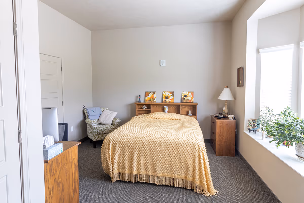 A bright bedroom with a bed covered by a yellow textured blanket, an armchair, nightstands, and a large window with plants.