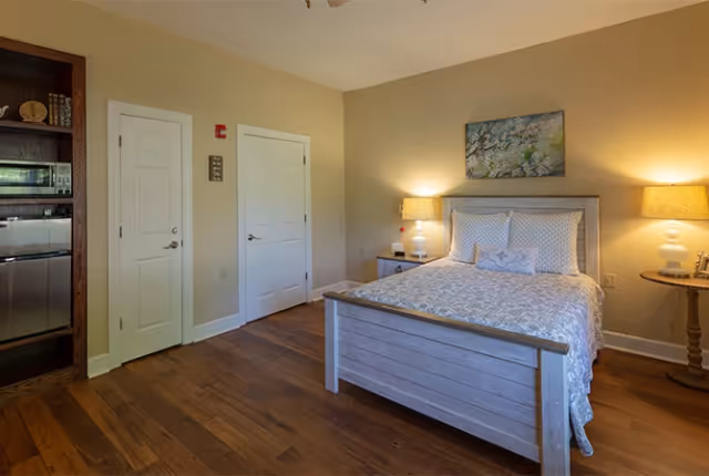 A cozy bedroom in a senior living facility featuring a wooden bed with a patterned bedspread and pillows, two bedside tables each with a lamp, a painting above the bed, wooden flooring, and two closed white doors on the adjacent wall. A small kitchenette area with a microwave and mini fridge is partially visible on the left side.