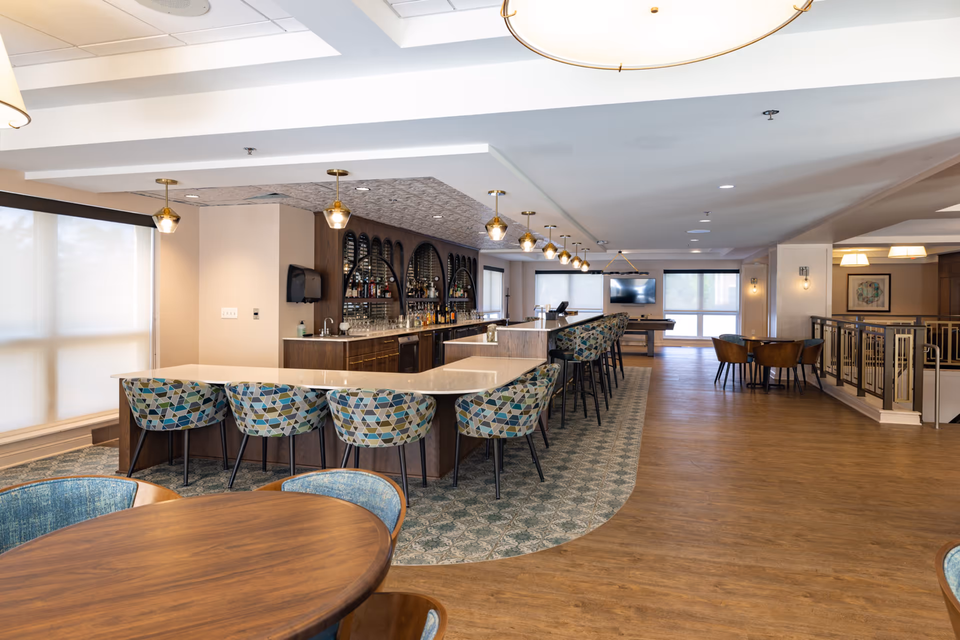 A spacious and modern common area featuring a long bar with patterned upholstered bar stools, pendant lighting, and a variety of bottles and glasses behind the bar. There are round wooden tables with chairs in the foreground and a pool table with a TV mounted on the wall in the background. The floor combines wood and patterned carpet, and large windows with shades allow natural light to fill the room.