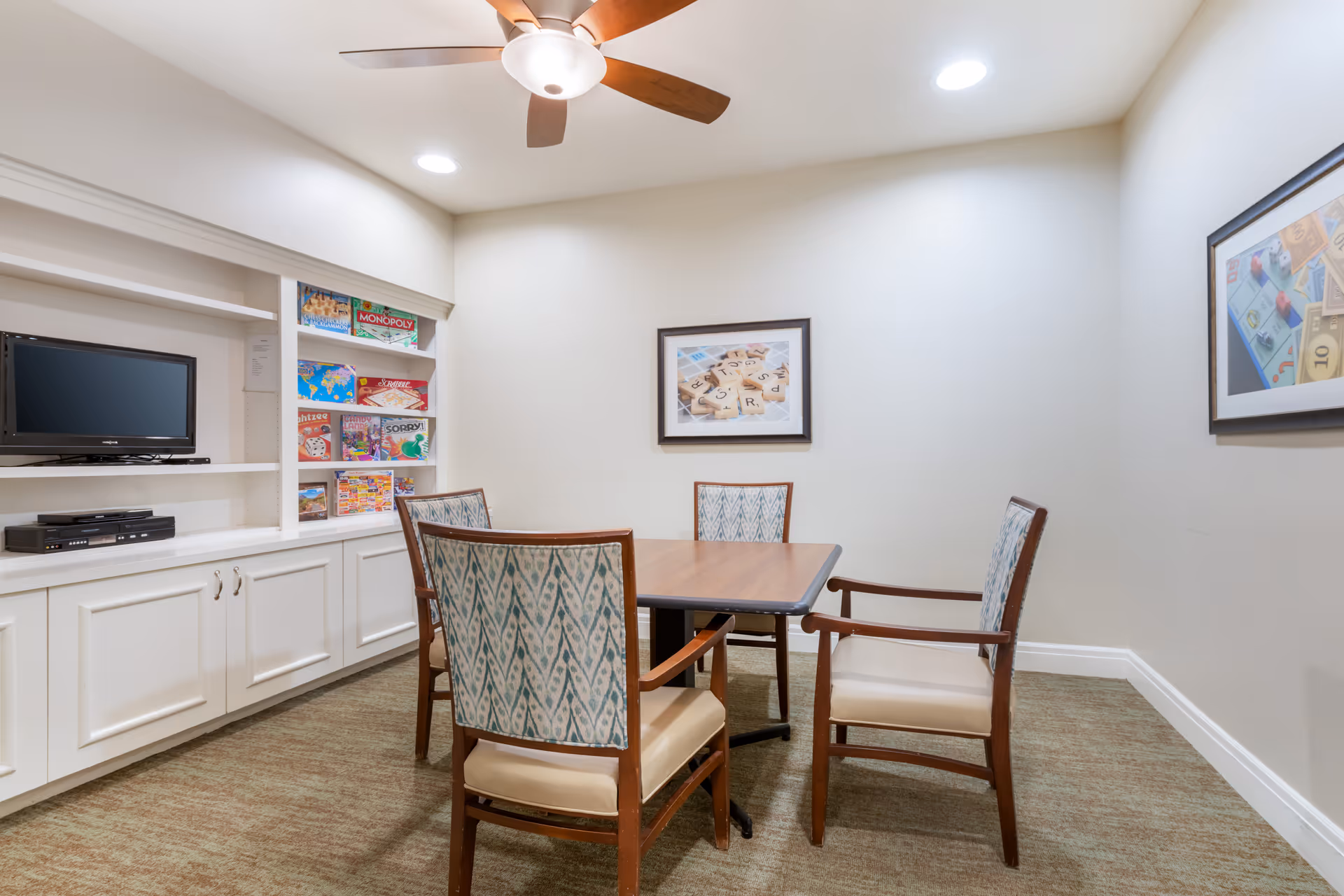 A small interior activity room with a square table surrounded by four chairs, built-in shelves holding board games and a TV, and framed artwork on the walls.