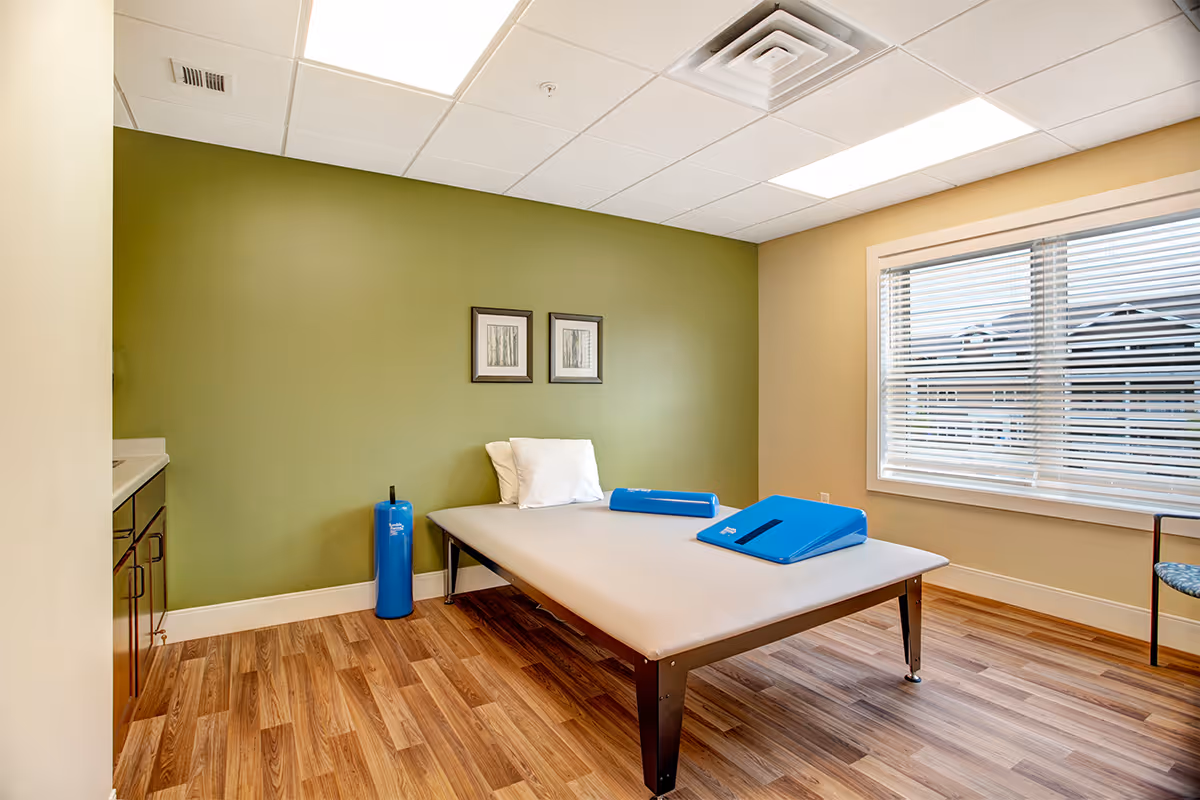 A small treatment room with a padded therapy table, green accent wall, window with blinds, and wood-look flooring.