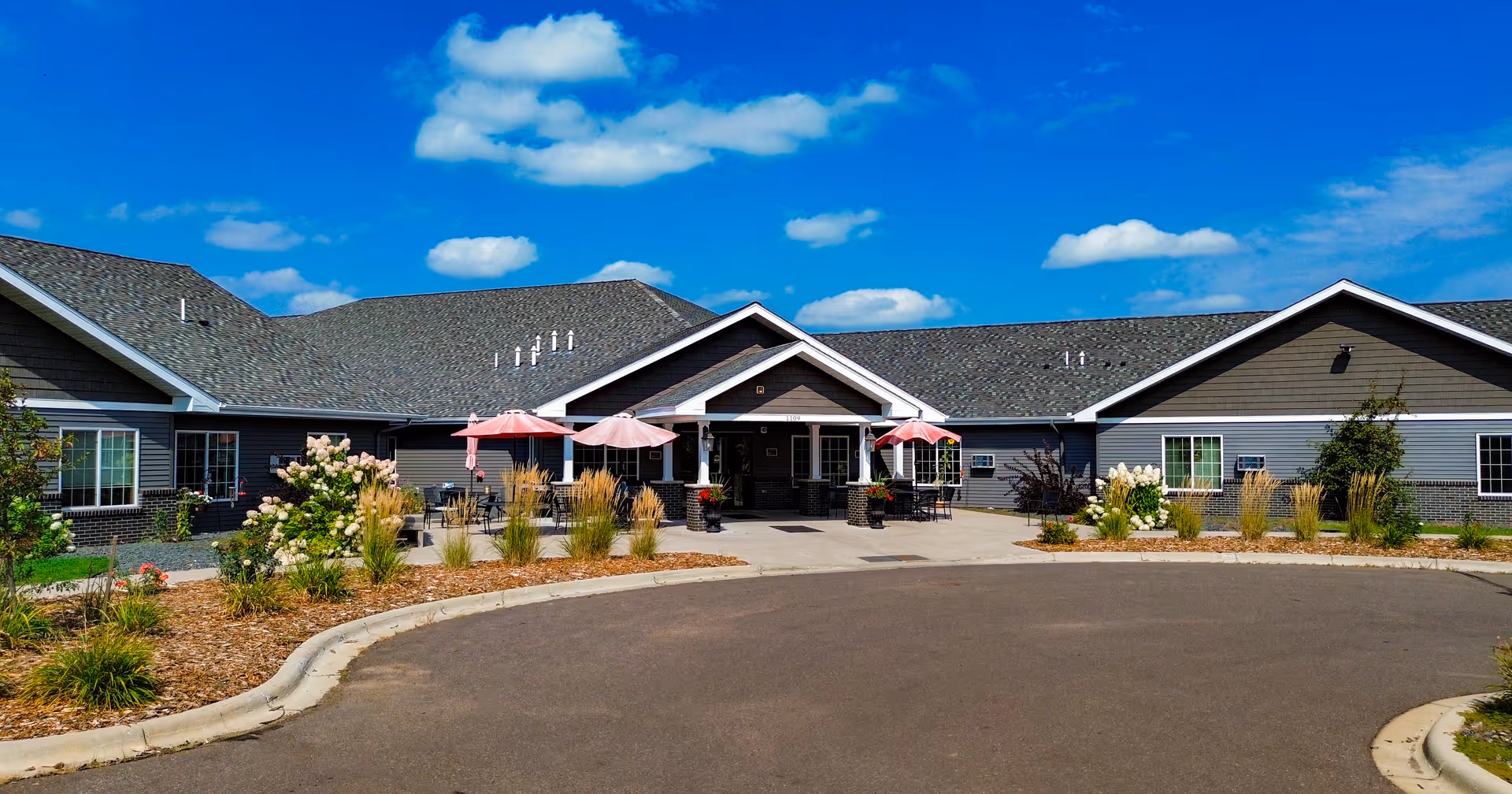 Front entrance of a single-story senior living building with outdoor seating and pink umbrellas and landscaped beds under a blue sky.