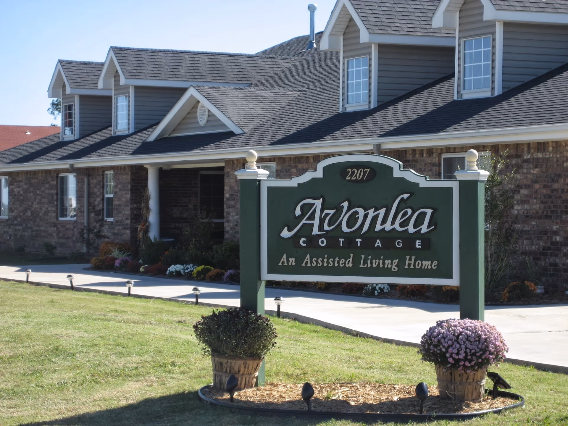 Exterior view of Avonlea Cottage, an assisted living home, showing a brick building with multiple windows and a gray shingled roof. In front of the building is a green and white sign with the name and address, surrounded by two flower pots with blooming flowers on a grassy lawn.