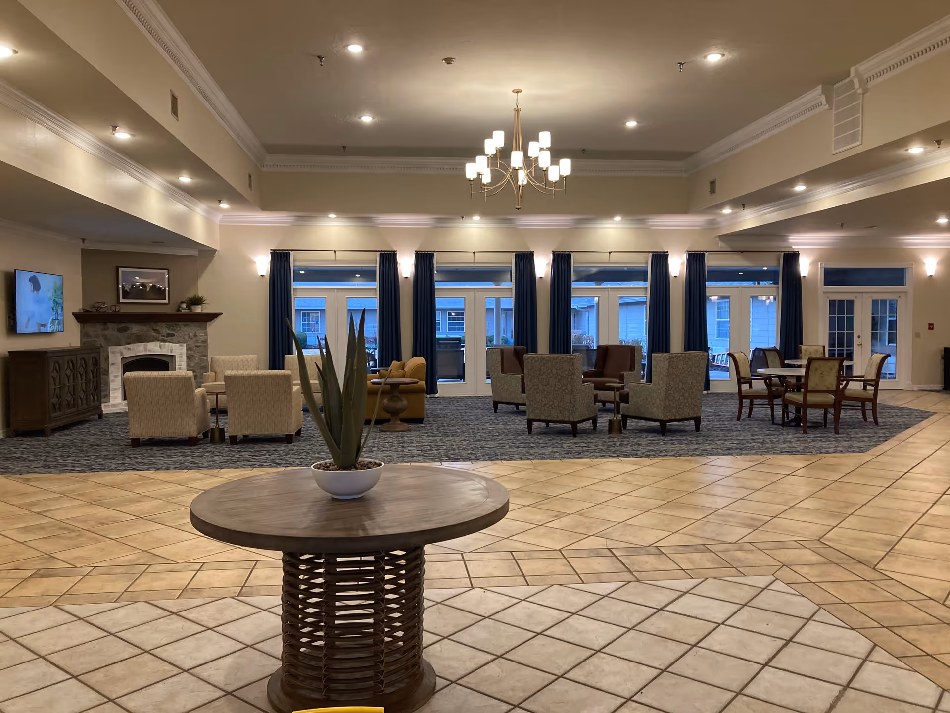 A spacious and well-lit common area with a tiled floor and a round wooden table with a potted plant in the foreground. In the background, there are multiple seating arrangements including armchairs and small tables on a carpeted area. Large windows with blue curtains and glass doors let in natural light. A chandelier hangs from the ceiling and a stone fireplace with a TV mounted above it is visible on the left side.