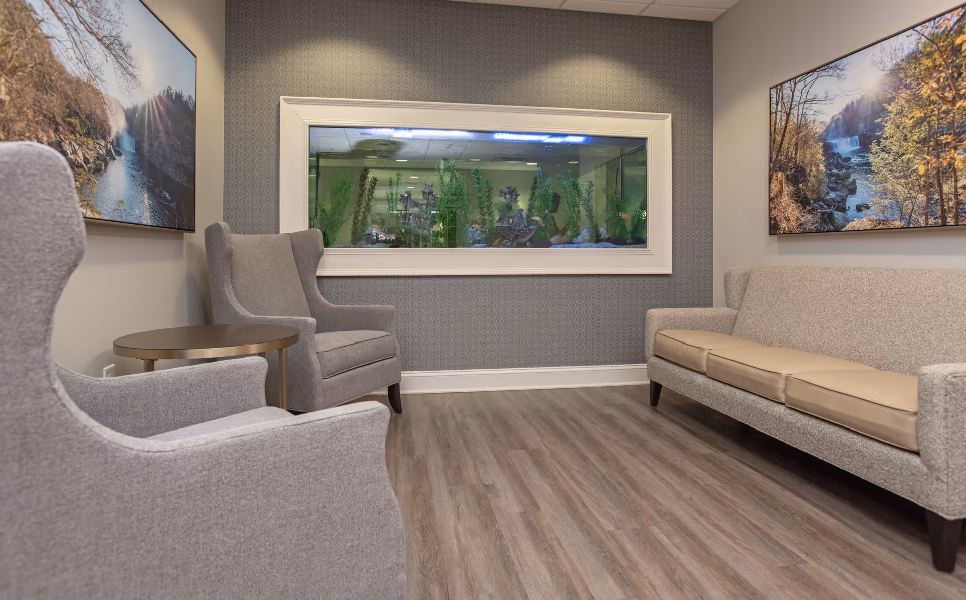 A cozy seating area with two gray armchairs and a gray sofa with beige cushions arranged around a small round wooden table. Behind the seating is a large built-in aquarium with green plants and decorative elements inside. The walls are decorated with two landscape paintings featuring nature scenes with trees and water. The floor is covered with wood-patterned vinyl or laminate.