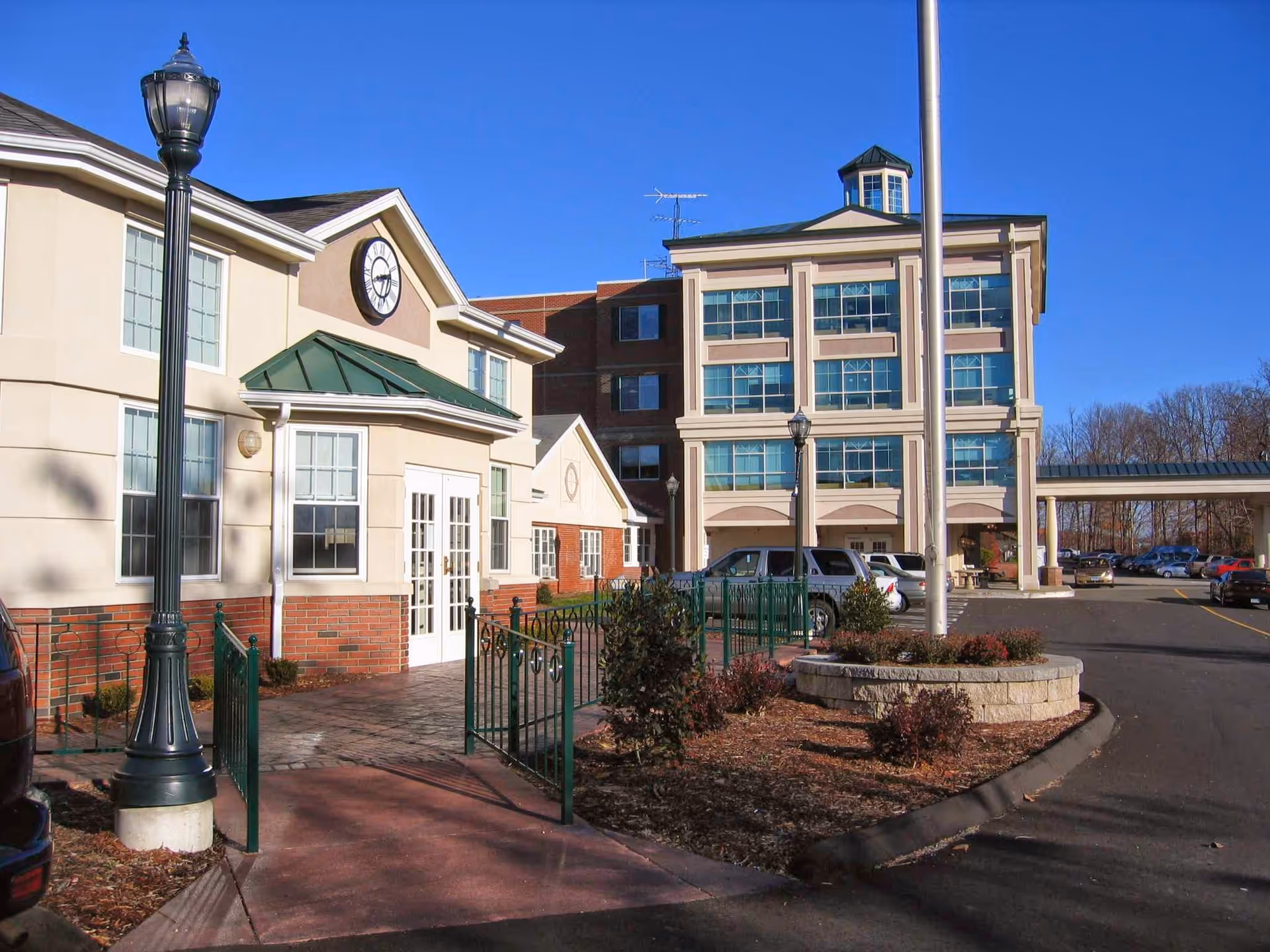 Front exterior of a multi-story nursing and rehabilitation building with an entrance, clock on the facade, lamp posts, flagpole, parked cars, and landscaped driveway under a clear blue sky.
