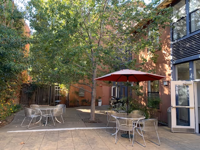 Outdoor patio area at Elders Inn with several metal tables and chairs, one table shaded by a red umbrella, surrounded by trees and adjacent to a building with large windows and an open door.