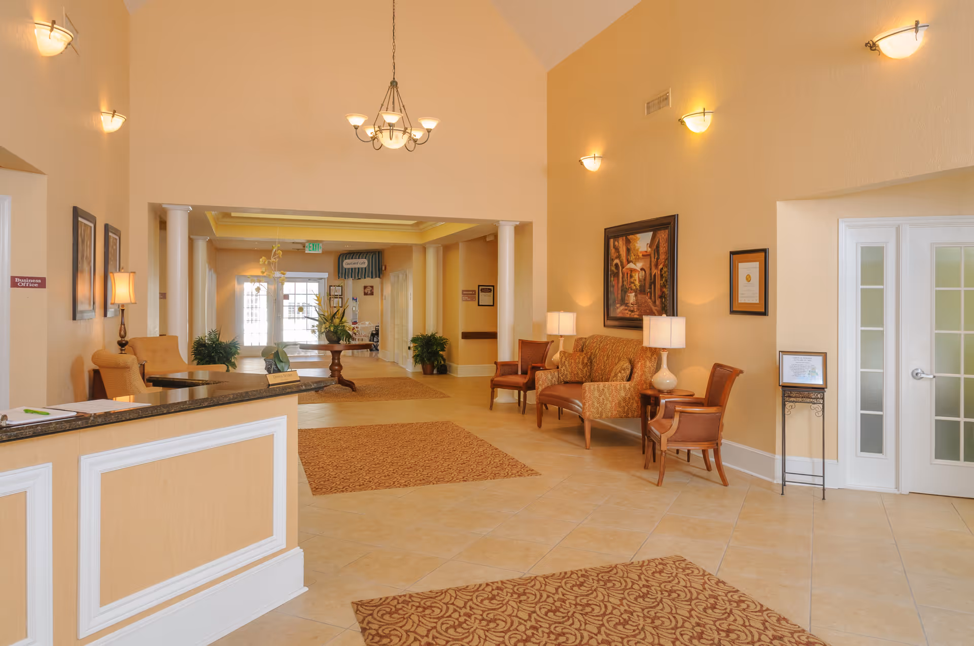 Reception area of a senior living facility with a front desk on the left, beige walls, tiled floor, patterned rugs, seating area with chairs and a couch, wall lamps, framed artwork, and a chandelier hanging from a high ceiling.