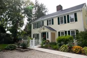 Exterior view of a two-story yellow house with green shutters, a small porch with a white railing, surrounded by trees and landscaped plants.