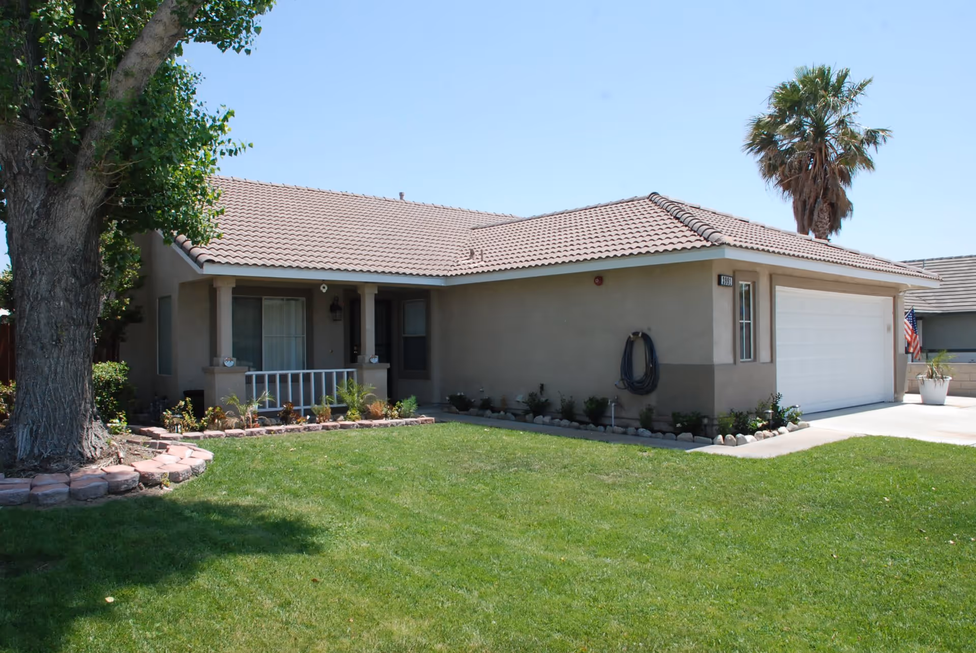 Single-story house with a tiled roof, beige exterior walls, a small front porch with white railing, a large green lawn, a tree on the left, and a palm tree near the right side of the house. There is a white garage door on the right and an American flag near the driveway.