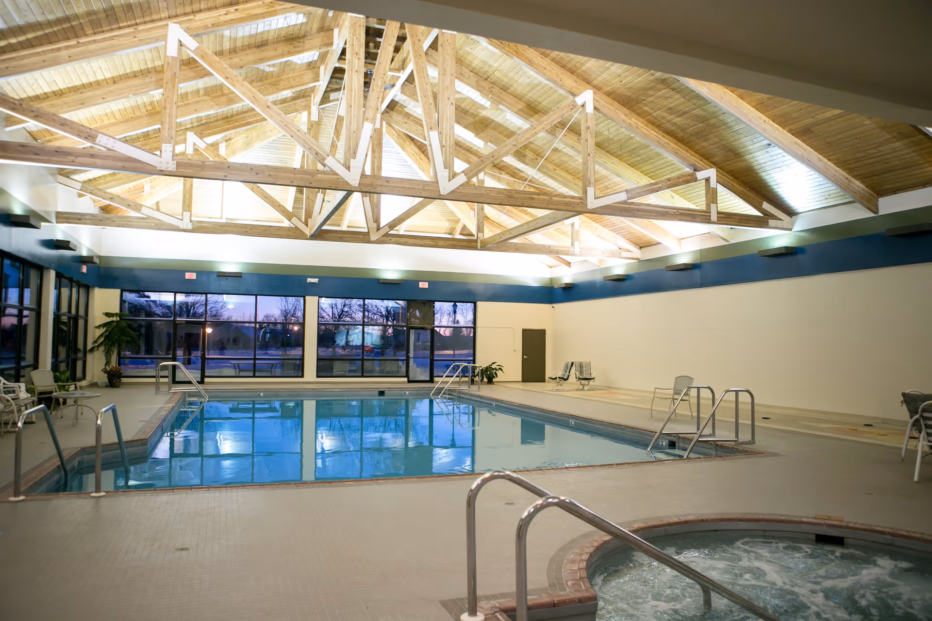 Indoor swimming pool area with a hot tub in the foreground, surrounded by chairs and tables. The ceiling features exposed wooden beams and skylights, and large windows line one side of the room showing an outdoor view at dusk.