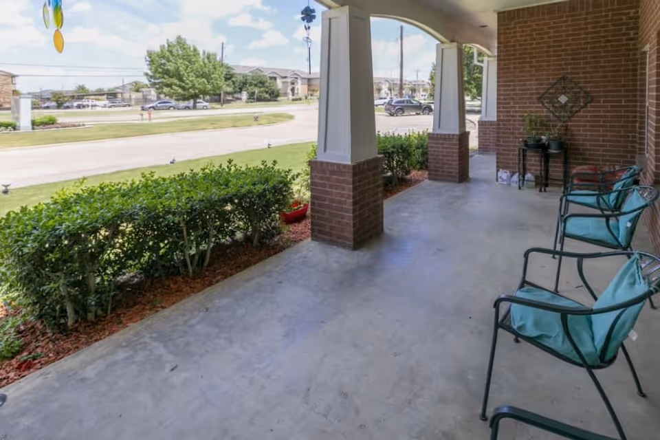Covered outdoor patio area with three metal chairs with teal cushions, brick pillars, and a small table with potted plants. Green bushes line the edge of the patio, and a street with cars and buildings is visible in the background under a partly cloudy sky.