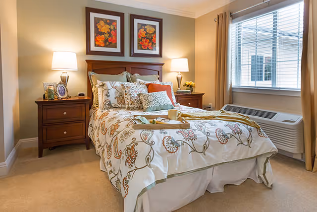 A cozy bedroom with a neatly made bed featuring floral patterned bedding and multiple pillows. There are two wooden nightstands on either side of the bed, each with a lamp and decorative items. Two framed floral artworks hang above the headboard. A window with beige curtains allows natural light into the room, and an air conditioning unit is positioned below the window.