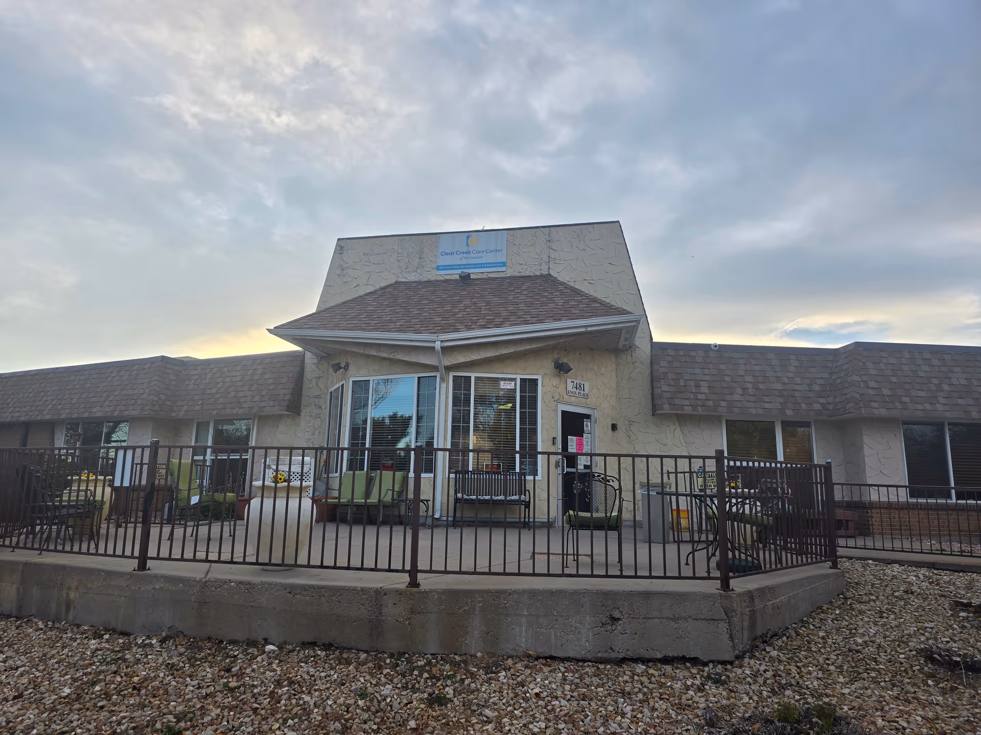 Exterior front entrance of Clear Creek Care Center with a fenced patio, seating, and a low stucco building under a cloudy sky.