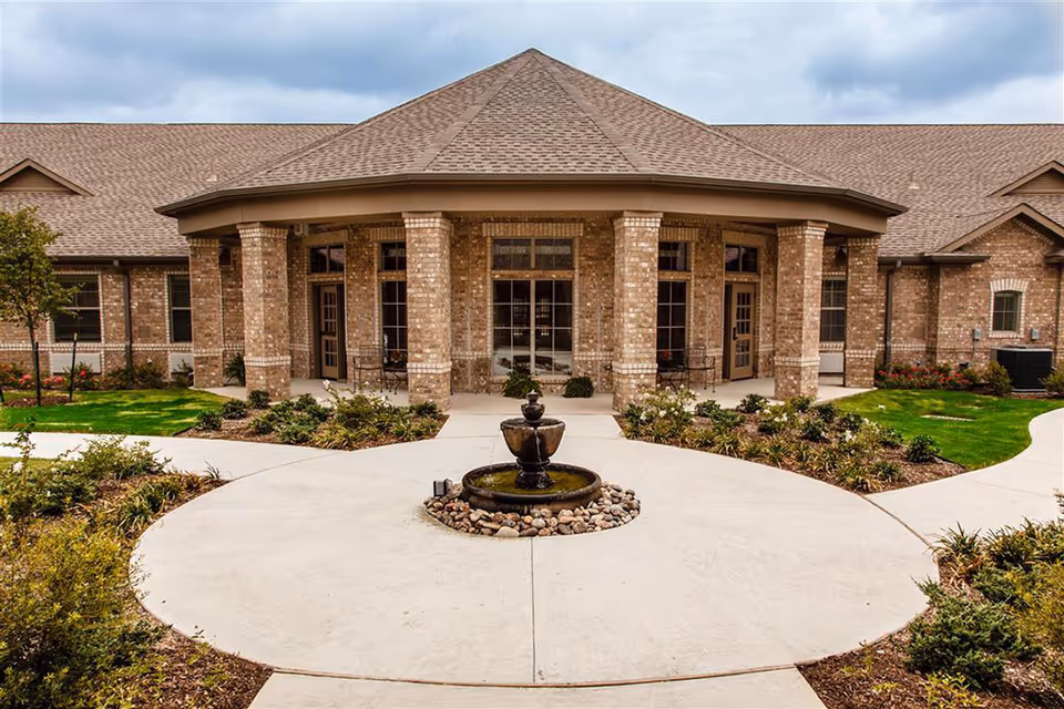 Front exterior view of The Harrison At Heritage facility showing a brick building with a covered entrance supported by columns, a circular driveway with a small fountain in the center, and landscaped greenery around the walkway.