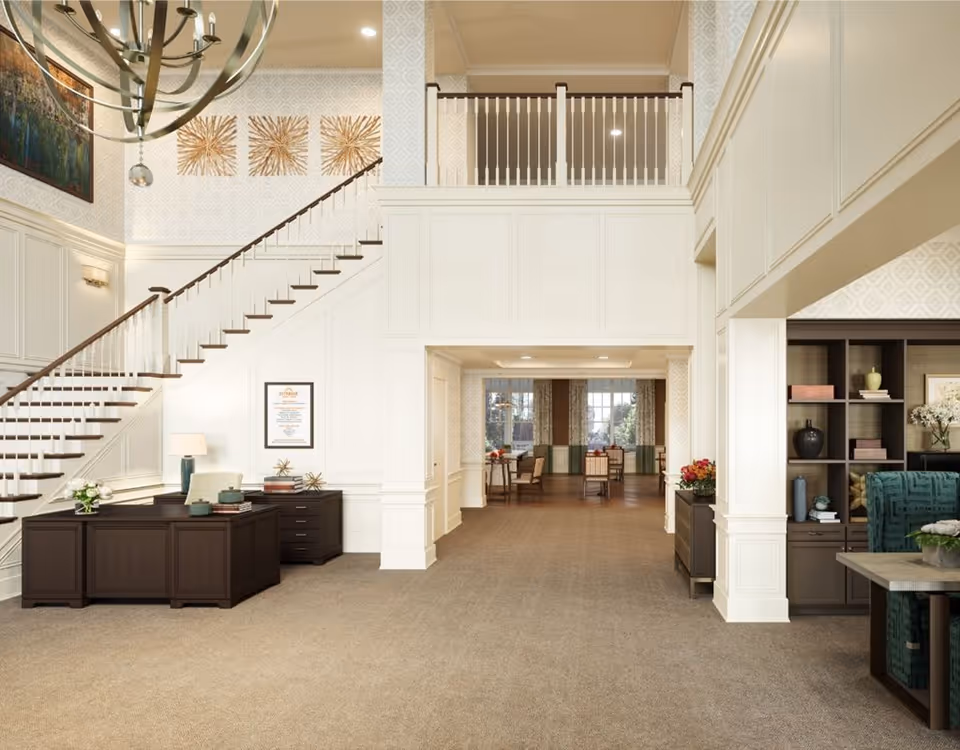 A spacious and elegant interior lobby area with a staircase on the left, a dark wooden reception desk with a lamp and decorative items, and a seating area with bookshelves and a green upholstered chair on the right. The walls are decorated with patterned wallpaper and artwork, and there is a chandelier hanging from the ceiling.