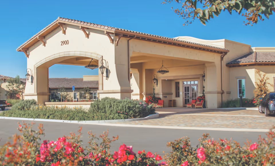 Exterior view of Creekview Health Center building entrance with a covered drop-off area, beige walls, and a tiled roof. There are decorative lantern-style lights hanging and mounted on the walls. The foreground shows a paved driveway and landscaped bushes and flowers.