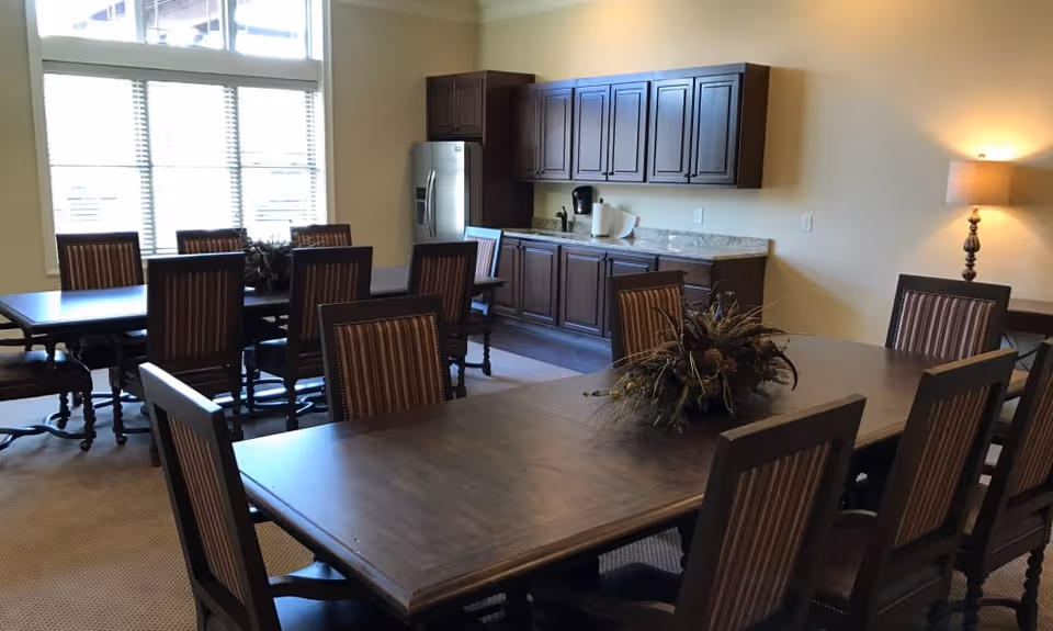 Interior view of a dining room with two large wooden tables surrounded by striped cushioned chairs. There is a decorative centerpiece on the nearest table. The room has large windows with blinds allowing natural light to enter. Against the far wall, there are dark wooden cabinets, a countertop with a coffee maker, and a stainless steel refrigerator. A lit table lamp is visible on the right side of the image.