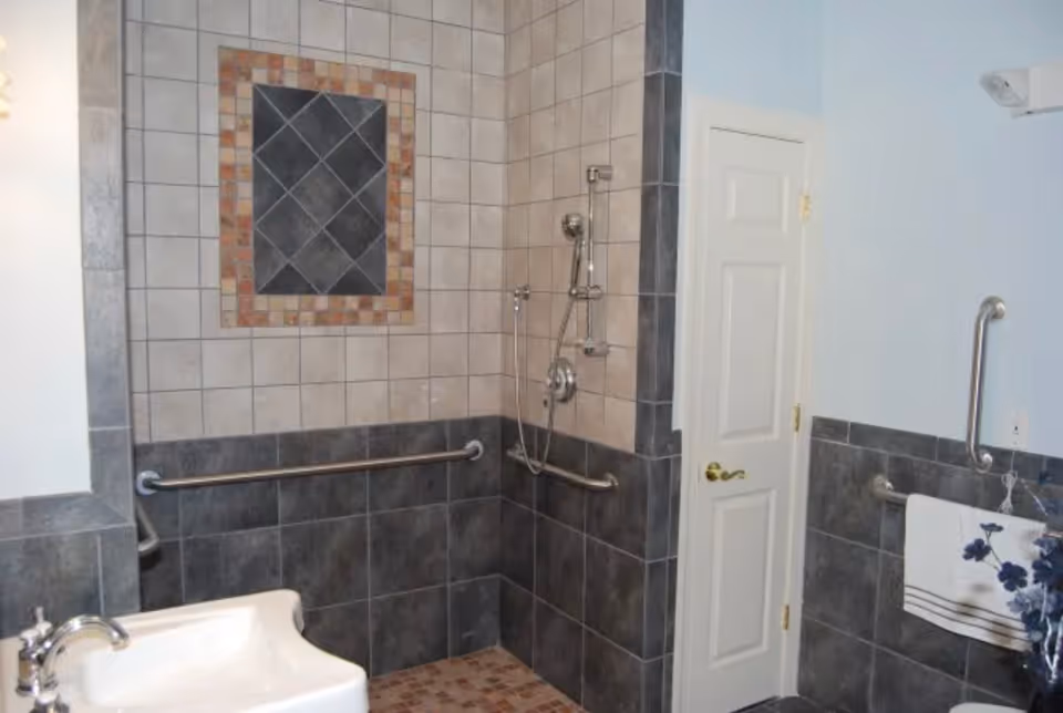 Accessible tiled bathroom with a walk-in shower, multiple grab bars, a sink, and a towel rack.