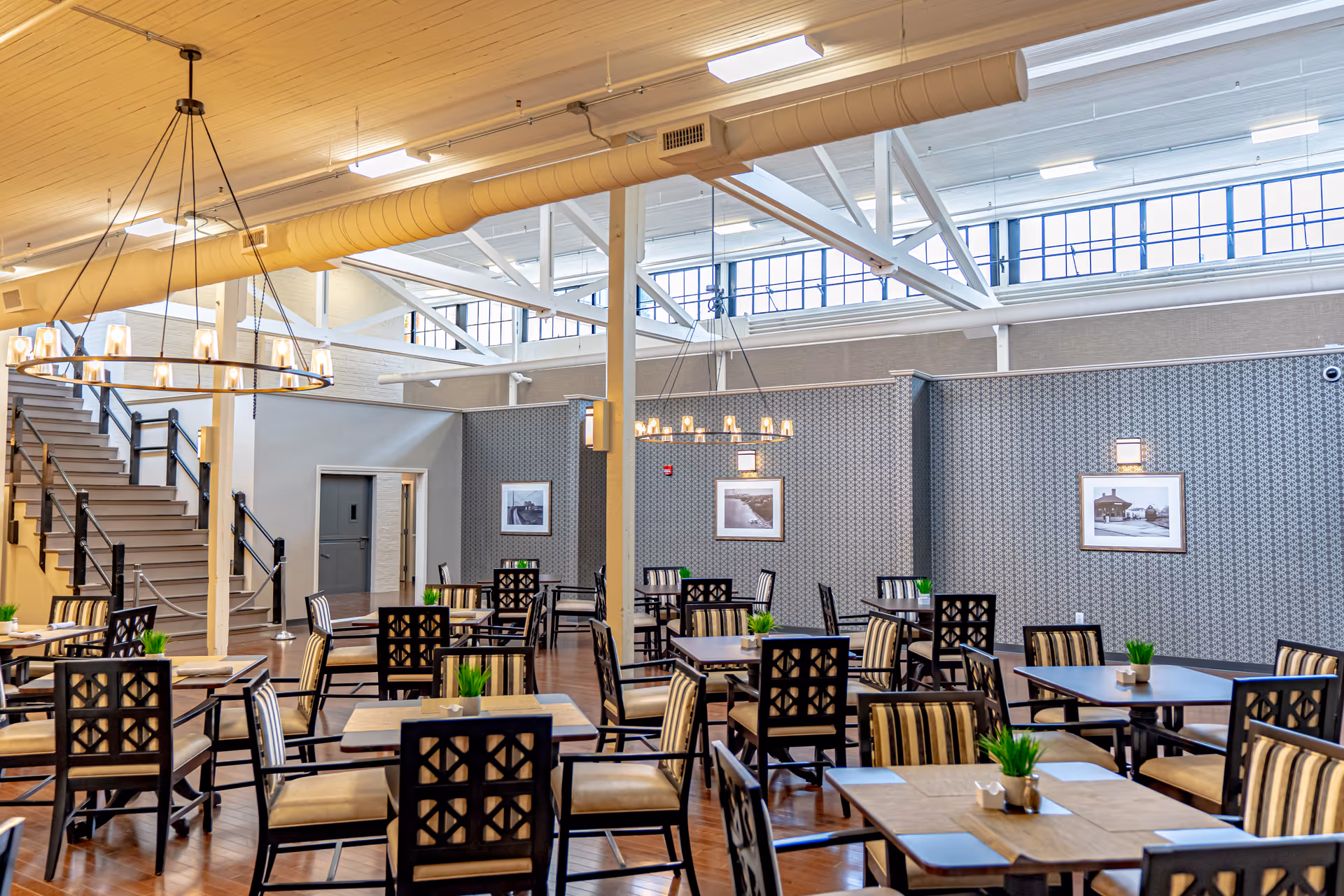A spacious dining room with multiple wooden tables and chairs arranged neatly. The chairs have patterned backs and striped cushions. Small green plants are placed on each table. The room features high ceilings with exposed beams and large windows near the ceiling allowing natural light to fill the space. Two modern chandeliers hang from the ceiling. There is a staircase on the left side and framed black and white photographs on the patterned wallpapered walls.