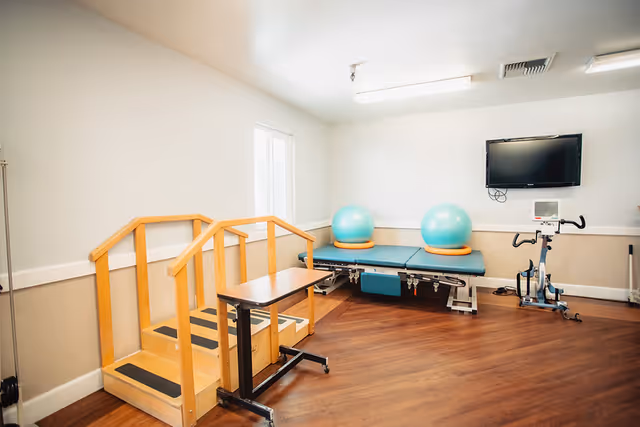 A physical therapy room with wooden stairs and handrails for exercise, two large blue exercise balls on padded tables, a stationary exercise bike, and a wall-mounted flat screen TV. The room has wood flooring and white walls with a window letting in natural light.