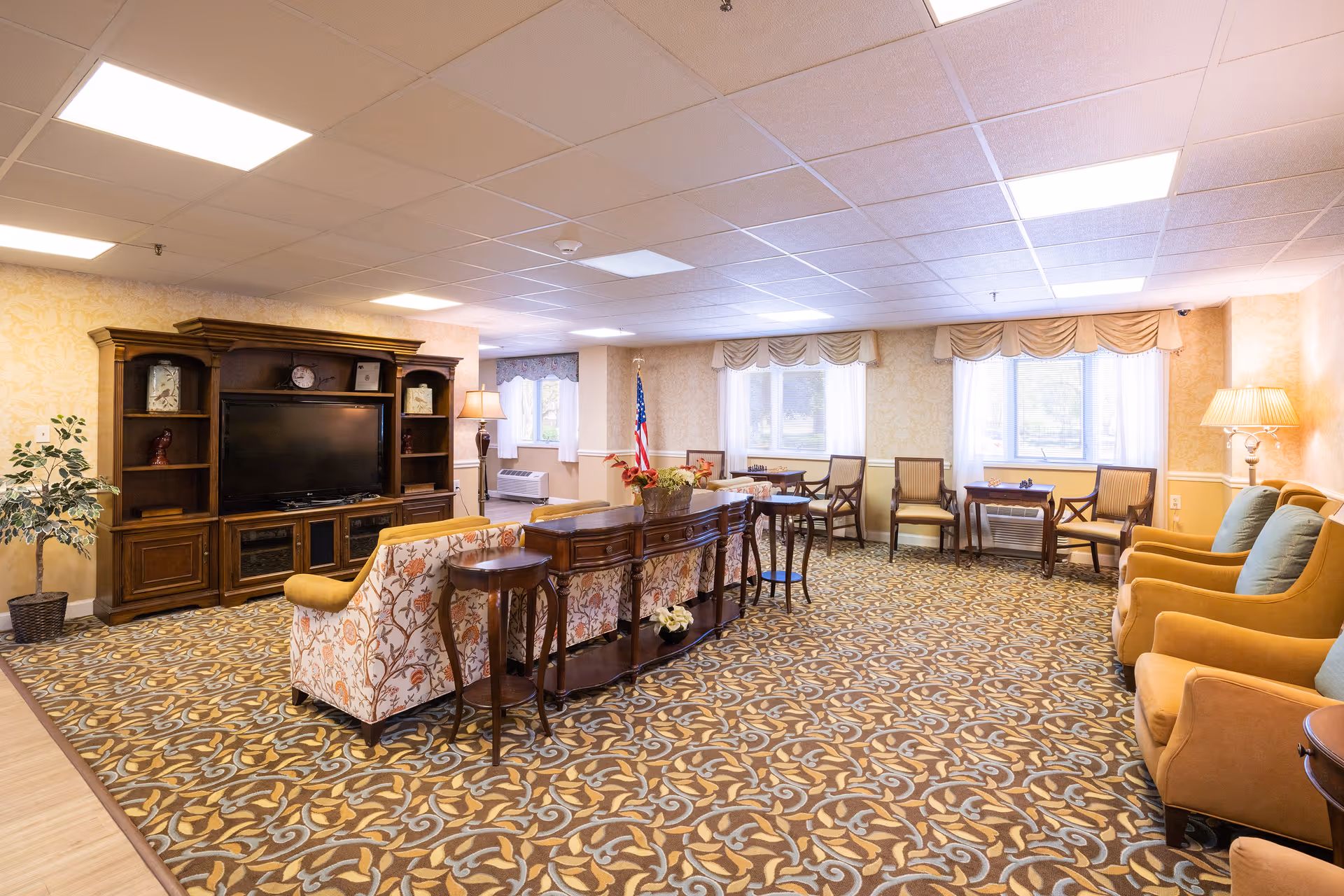 A spacious living room area in a senior living facility with patterned carpet, floral upholstered sofas, wooden side tables, and a large wooden entertainment center with a flat-screen TV. The room has multiple chairs arranged near windows with sheer curtains and valances, an American flag in the corner, and warm lighting from floor lamps.