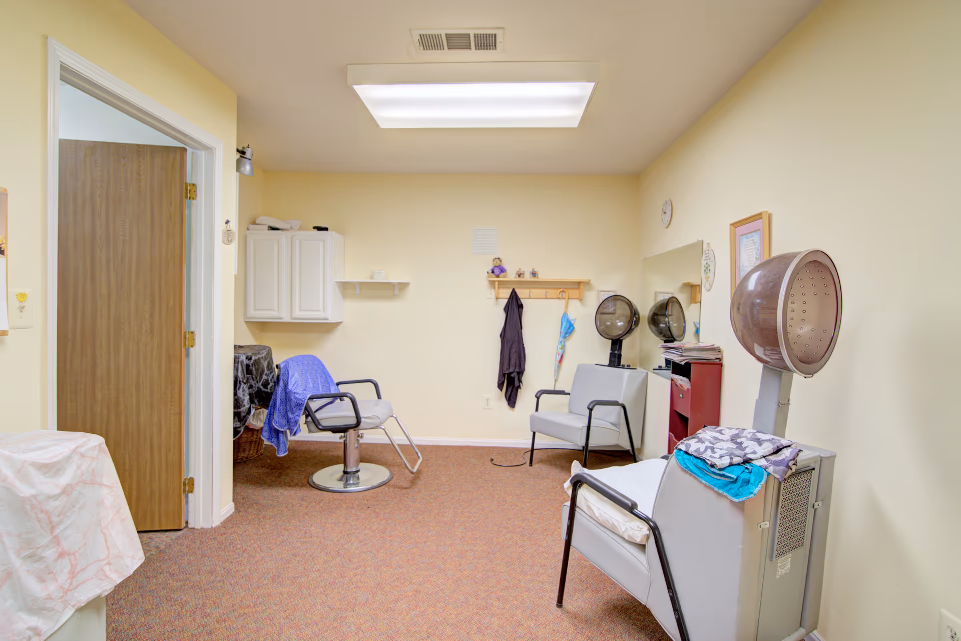 Small salon-style room with styling chairs, hooded hair dryers, cabinets and a coat rack in a senior living facility.
