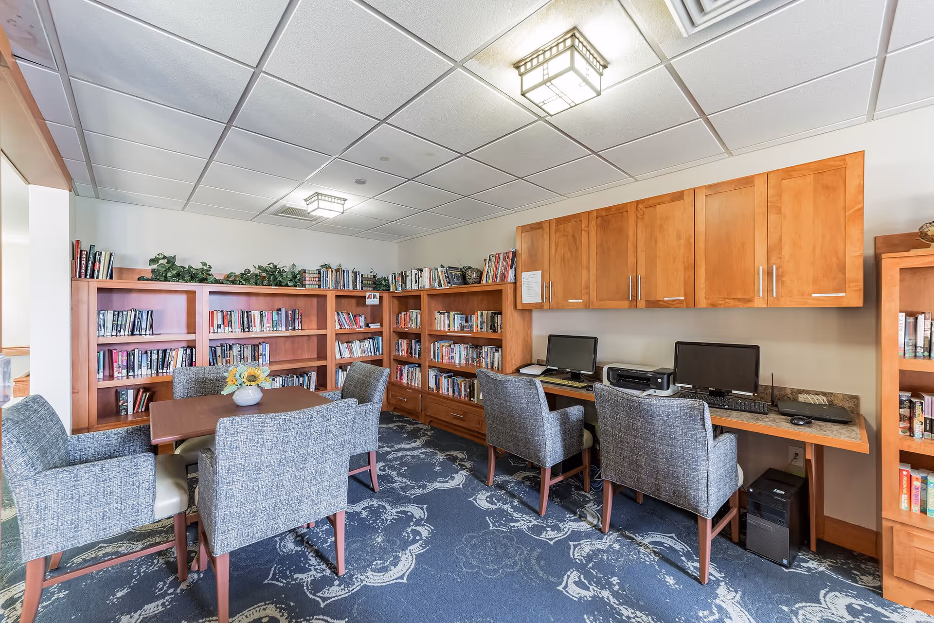 A cozy room with wooden bookshelves filled with books along the walls, a table with four upholstered chairs in the center, and a workspace with two computers and a printer under wooden cabinets. The room has a patterned carpet and a ceiling with recessed lighting.