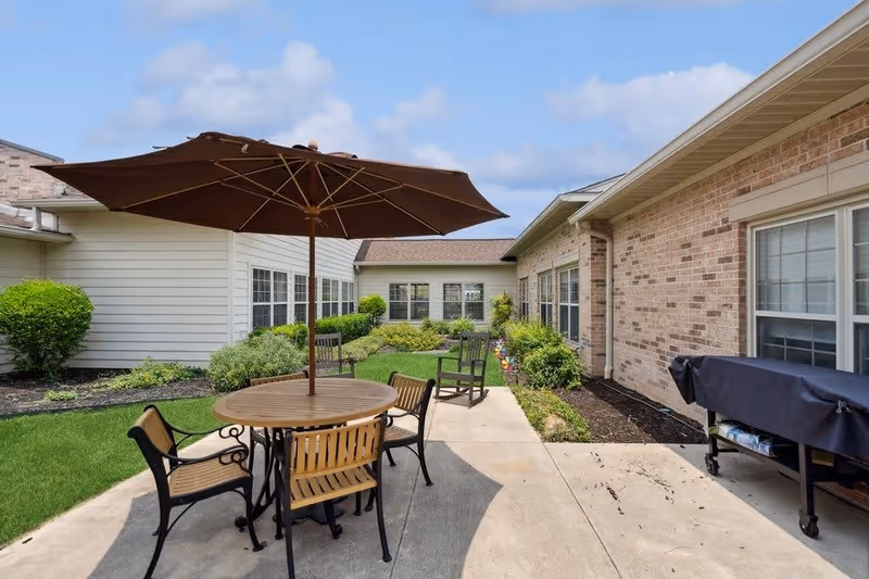 Outdoor patio area with a round wooden table and four chairs under a large brown umbrella. The patio is surrounded by a well-maintained garden with bushes and grass, adjacent to a brick and siding building with multiple windows.