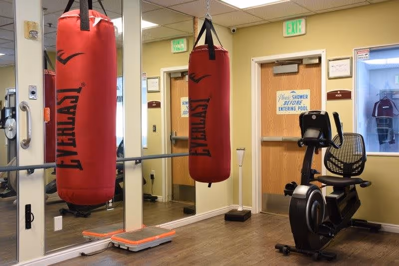 Indoor fitness room with a red Everlast punching bag hanging from the ceiling, a stationary exercise bike, a scale on the floor, and mirrored walls reflecting the equipment and doors. There are two wooden doors, one with a sign that reads 'Please SHOWER BEFORE ENTERING POOL.'