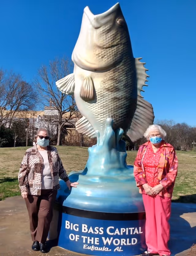 Two elderly women wearing face masks standing on either side of a large fish statue with a blue base that reads 'Big Bass Capital of the World, Eufaula, AL' in an outdoor park setting with trees and a clear blue sky.