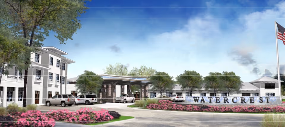 Exterior view of Watercrest Santa Rosa Beach senior living facility showing a large white multi-story building with several parked cars in front, landscaped greenery and pink flowers, and an American flag on a flagpole to the right.