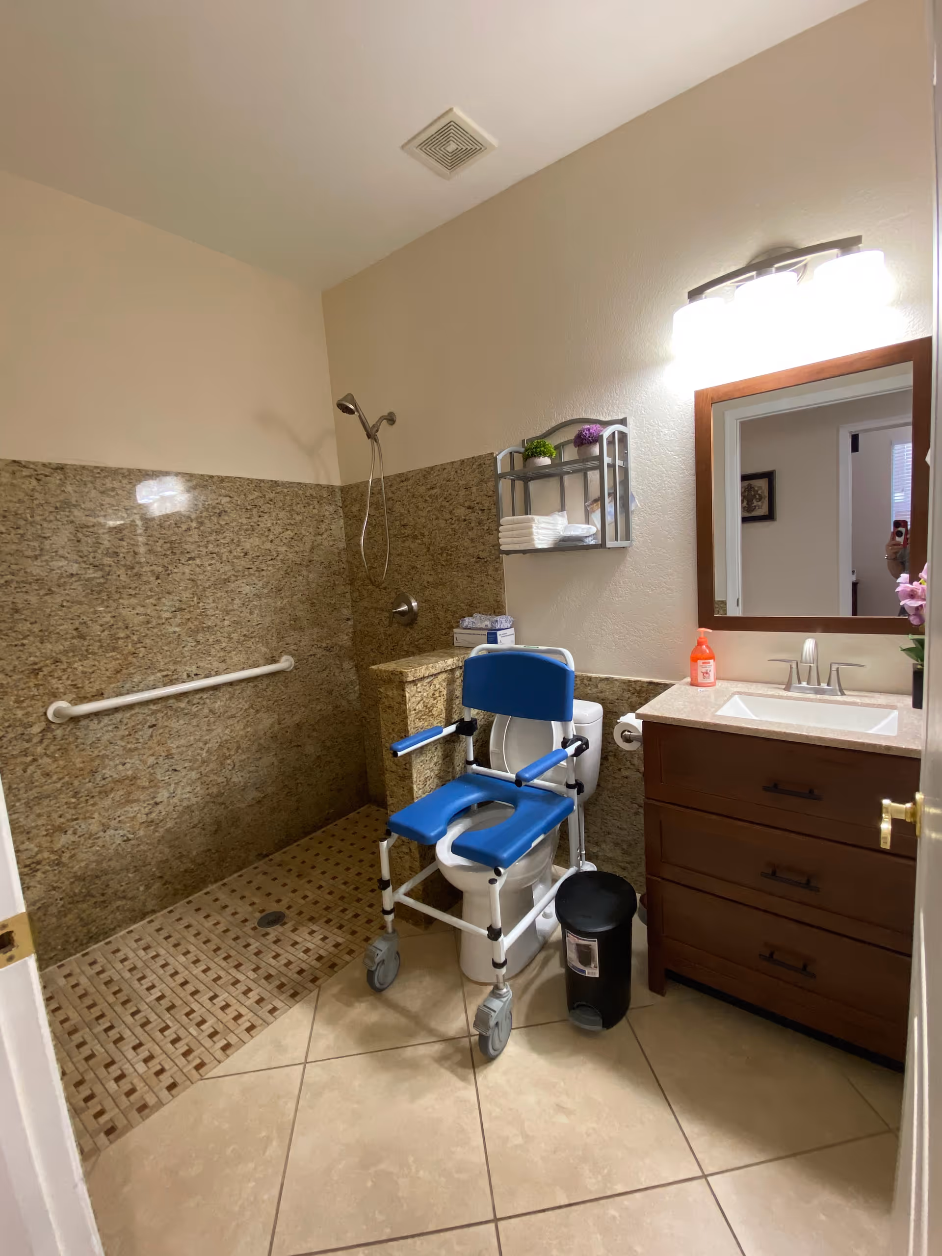 Accessible bathroom with a roll-in shower, grab bar, wheeled commode chair, sink vanity and mirror.