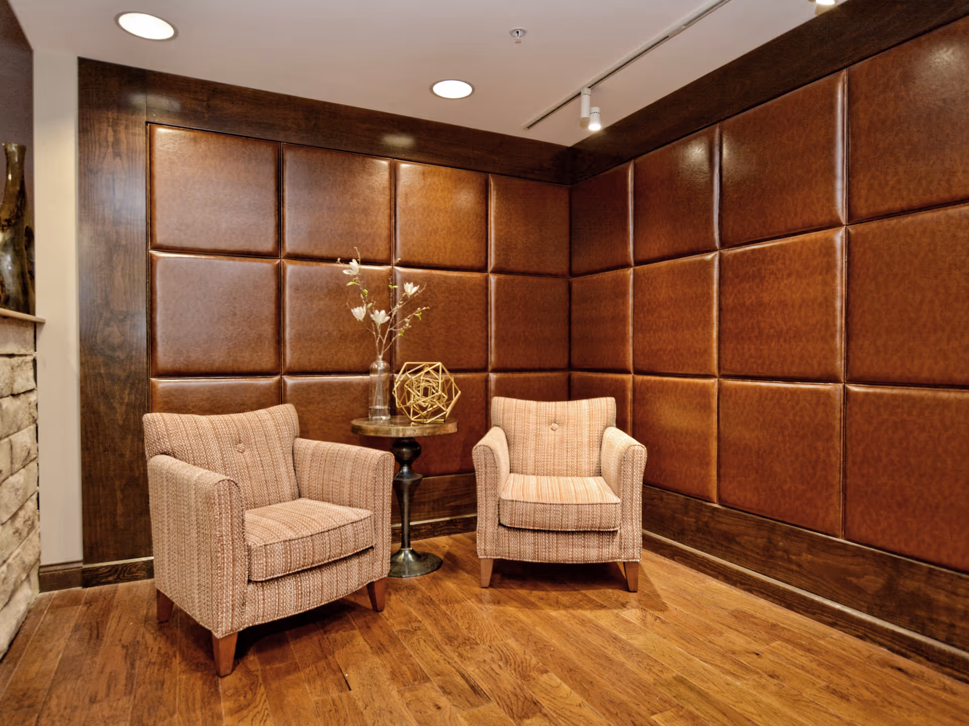 Two upholstered armchairs flank a small round table in a cozy corner with padded leather wall panels and hardwood flooring.