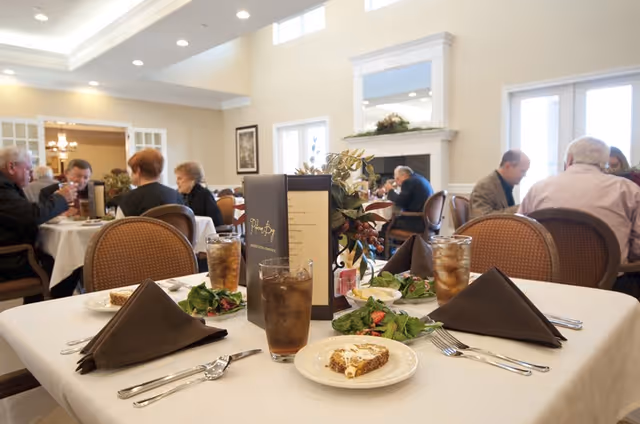 A table set for a meal with salads, iced tea, and a dessert in a bright assisted-living dining room while residents dine in the background.