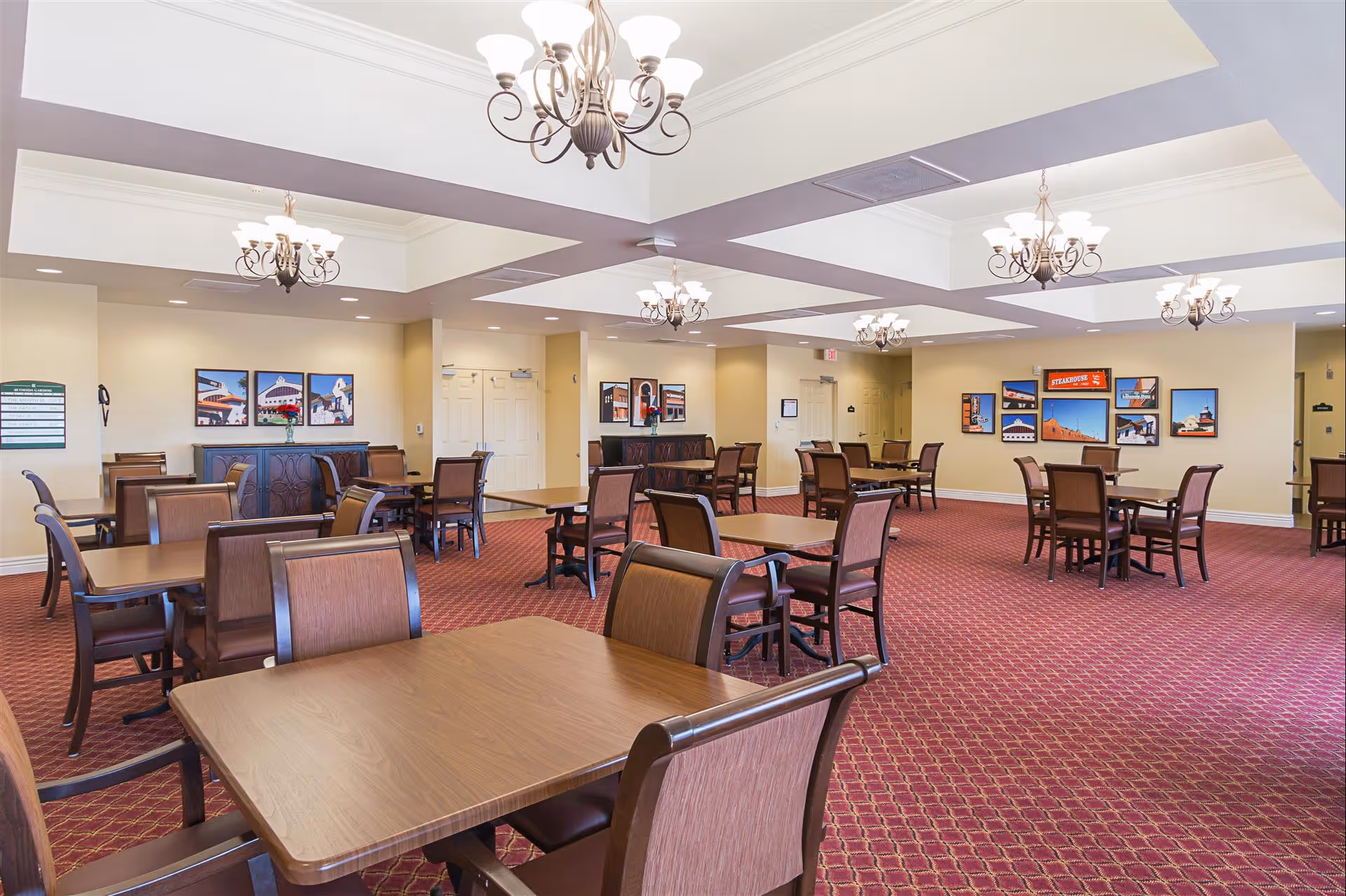 A spacious dining room with multiple wooden tables and chairs arranged neatly on a red patterned carpet. The ceiling features several elegant chandeliers providing warm lighting. The walls are decorated with framed pictures and there are double doors and an exit door visible in the background.