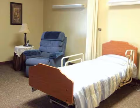 Hospital-style bedroom featuring a bed, blue recliner, side table with lamp, and framed art.