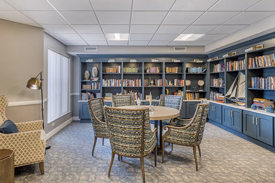 A cozy library or reading room with a round wooden table surrounded by six patterned chairs. The walls are lined with blue built-in bookshelves filled with books and decorative items, including a model sailboat. There is a beige armchair with a blue cushion and a floor lamp near a window with vertical blinds. The room has a carpeted floor and a white tiled ceiling with recessed lighting.