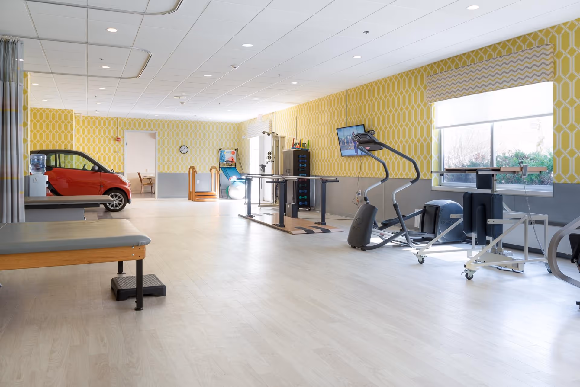 Bright rehabilitation/exercise room with cardio machines, therapy benches, and a small red practice car near the back.