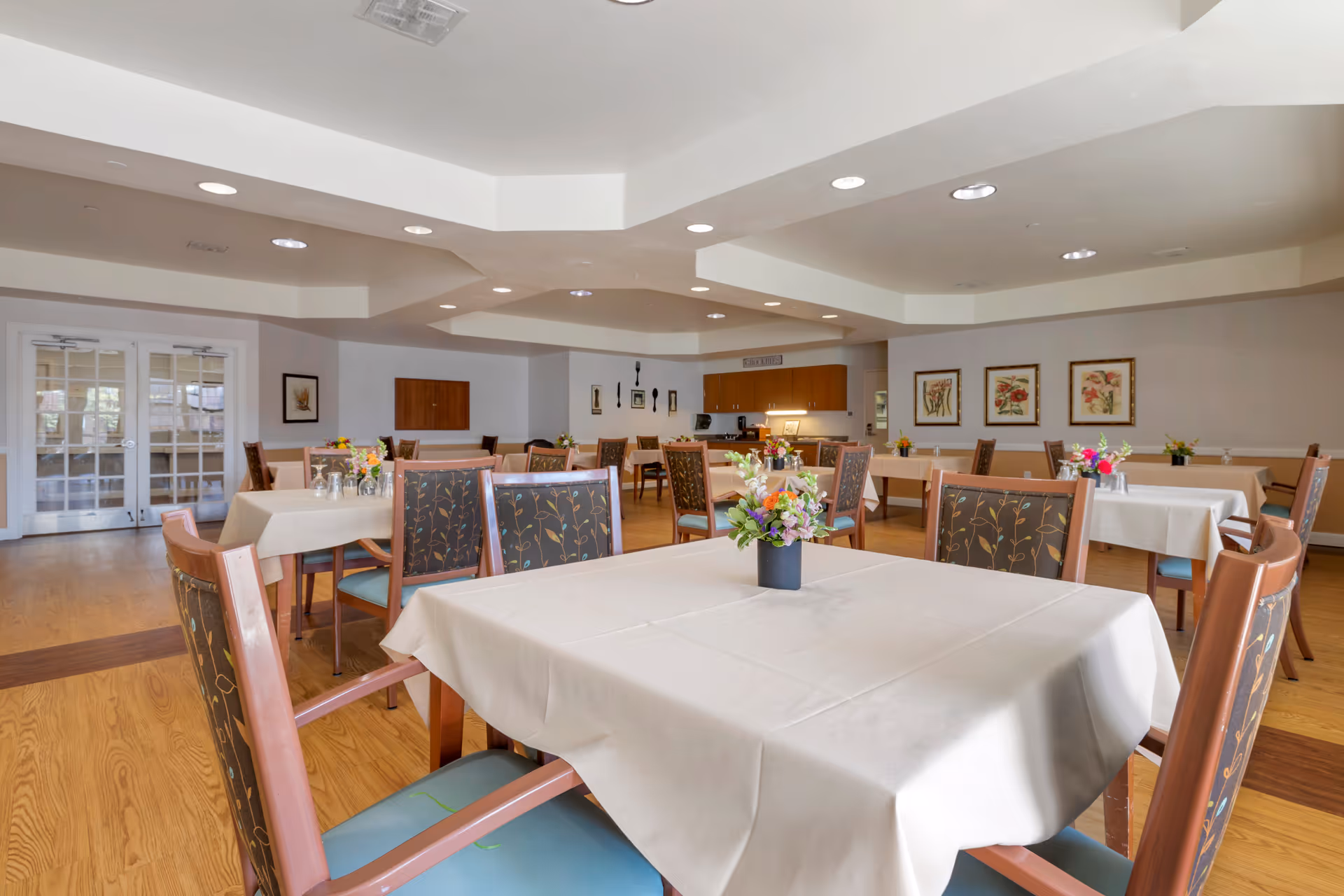 A spacious dining room with multiple tables covered in white tablecloths, each adorned with a small floral centerpiece. The room has wooden floors, cushioned chairs with floral patterned backs, framed artwork on the walls, and recessed lighting in the ceiling. Double glass doors are visible on the left side of the room.